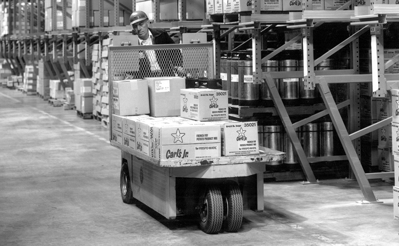 Black and white image of a man driving a stock cart loaded with Carl's Jr. boxes
