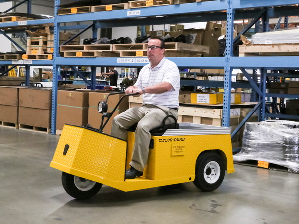 Man riding a yellow utility cart through a warehouse full of cardboard boxes